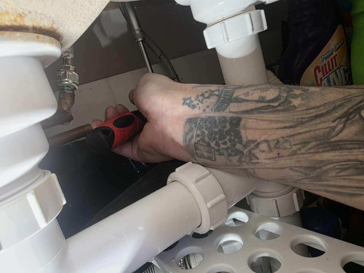 Plumber working on pipework under a kitchen sink in a tight cabinet.